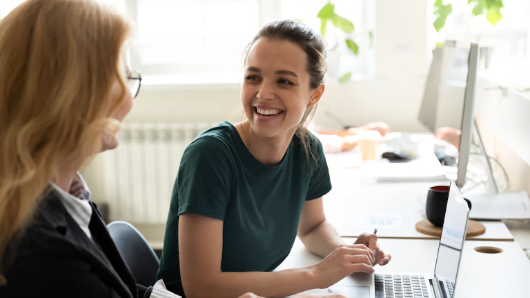 Pleasant workday two women colleagues employees diverse age sharing points view workplace office happy smiling millennial female trainee discussing business ideas with adult aged trainer